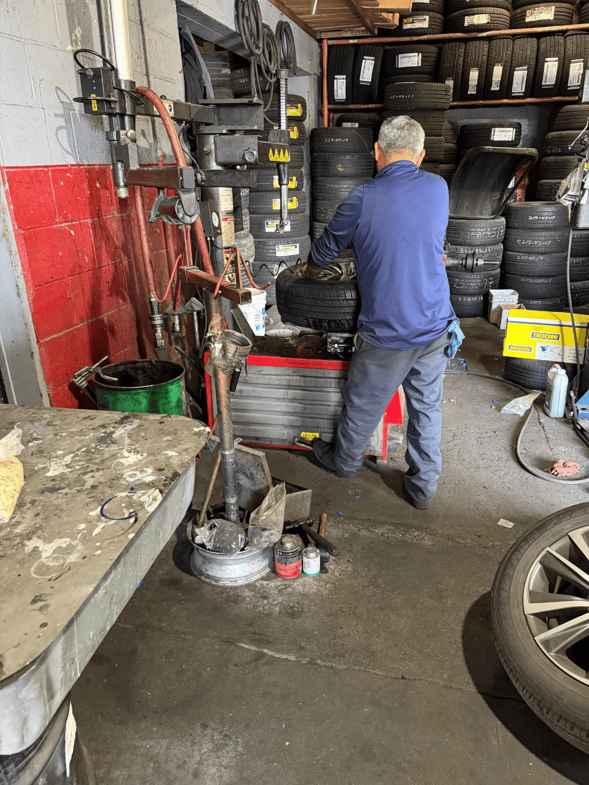 Tire technician inspecting a tire at a tire shop, surrounded by stacked tires and tire-changing equi.